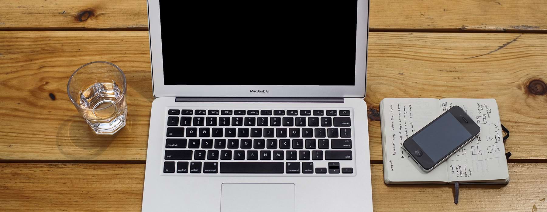 over head shot of open laptop on wooden desk with glass of water, notebook and cell phone